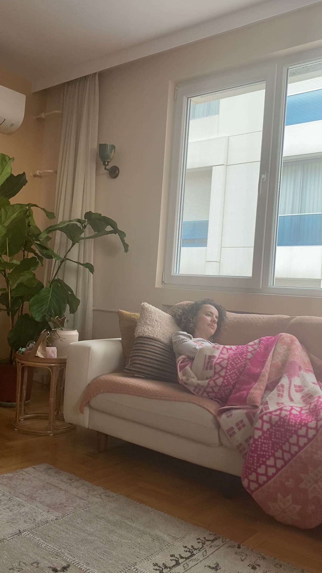 A woman lying on a sofa under a blanket, looking out of the window